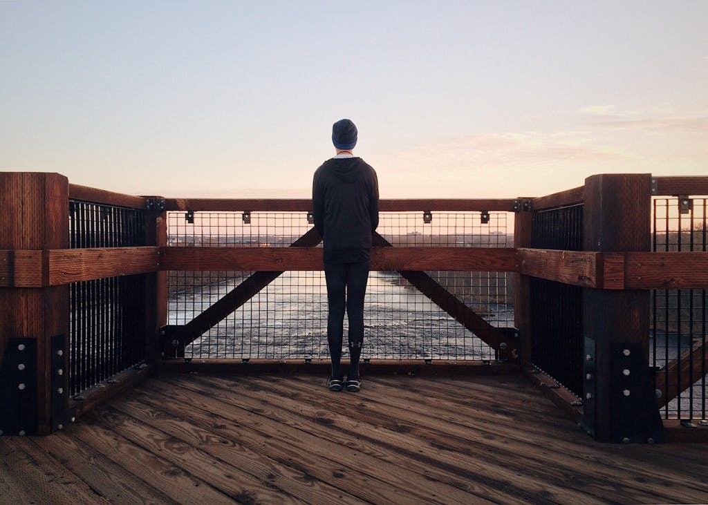 A person stands alone on a wooden bridge overlooking a serene river at sunset.